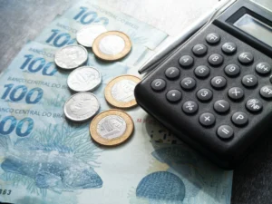 Cédulas e moedas de reais ao lado de uma calculadora, representando análise de custos fixos e variáveis em uma empresa.