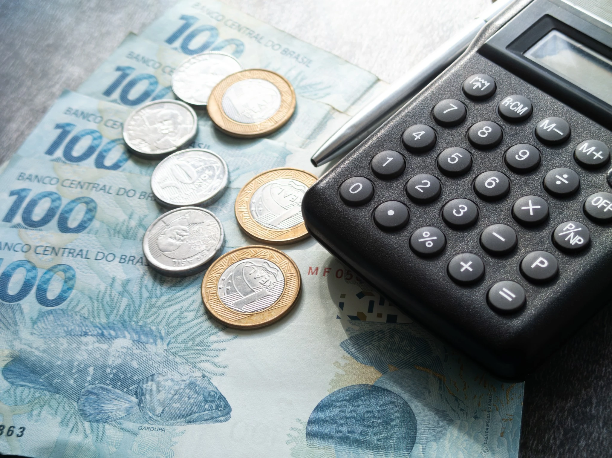 Cédulas e moedas de reais ao lado de uma calculadora, representando análise de custos fixos e variáveis em uma empresa.